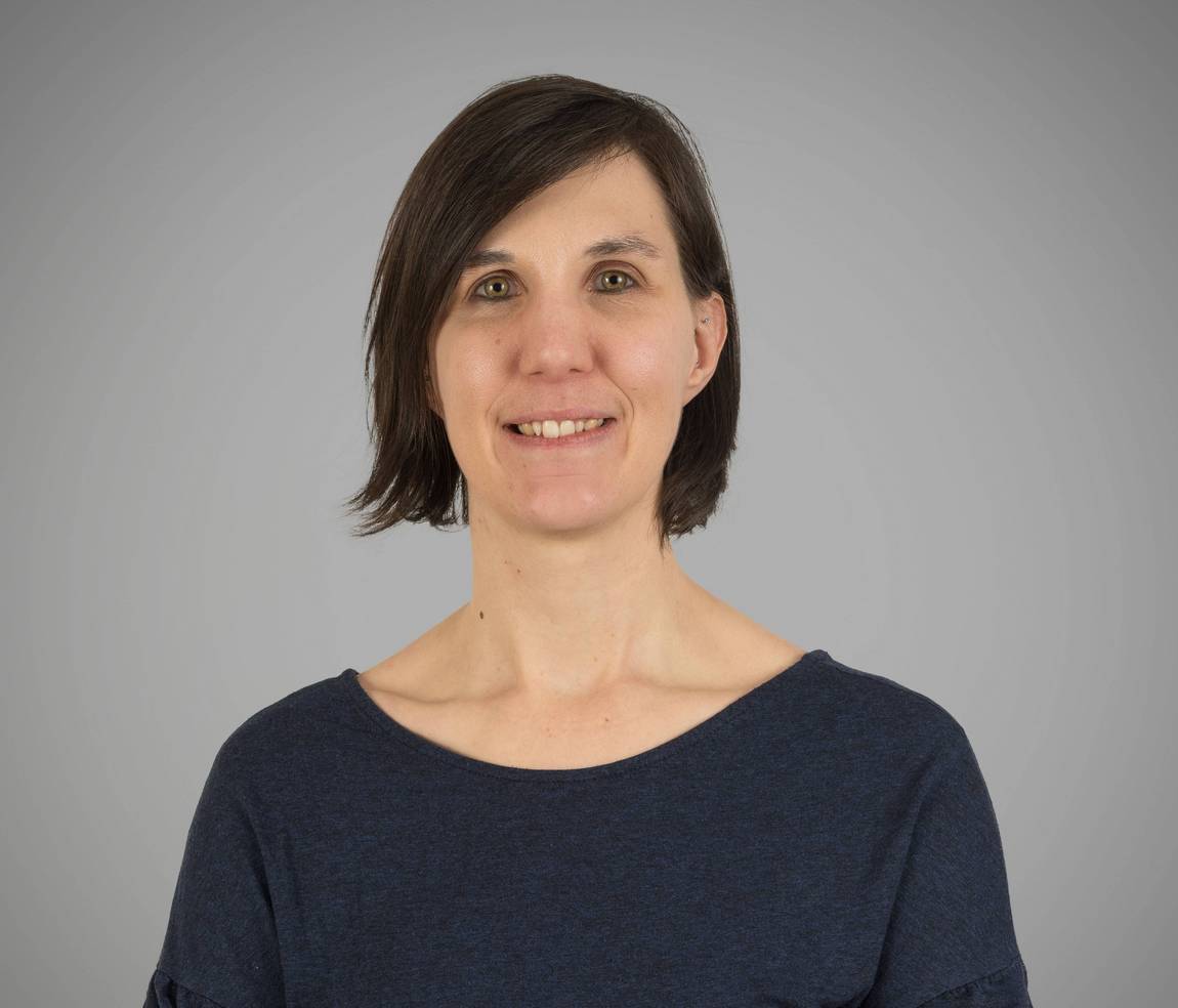 Eine Frau mit glattem braunem Haar und einem leichten Lächeln, die ein dunkelblaues Oberteil trägt, steht vor einem schlichten hellgrauen Hintergrund.